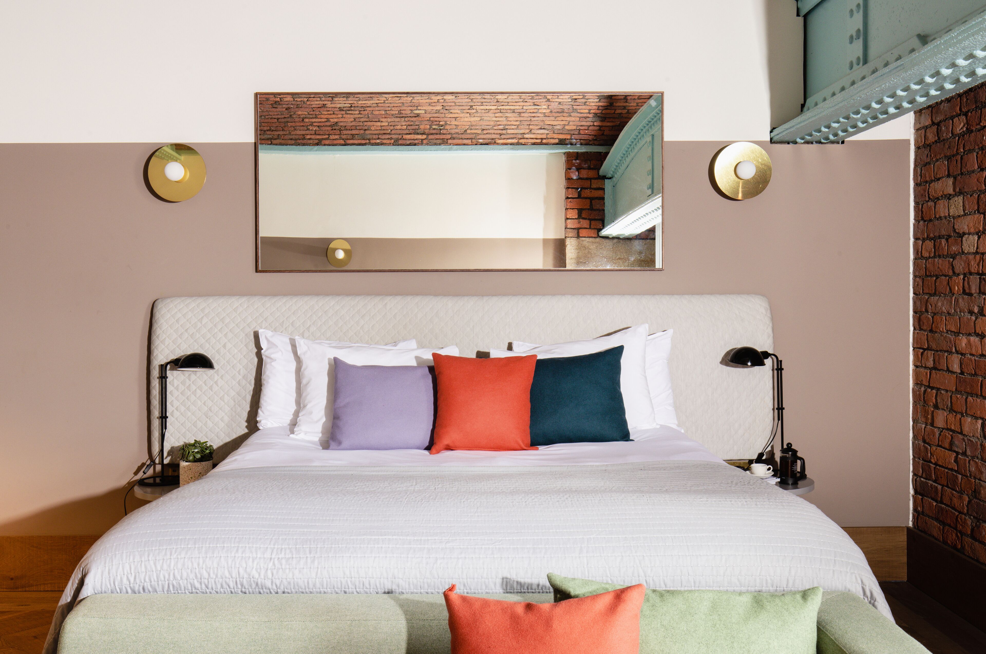 A neatly made bed with a white headboard, multiple pillows in various colors, two bedside lamps, and a small plant on the nightstand.