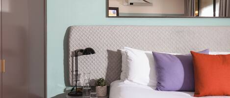 A hotel room with a bed, a bedside table with a lamp, a small plant, and a mirror on the wall.