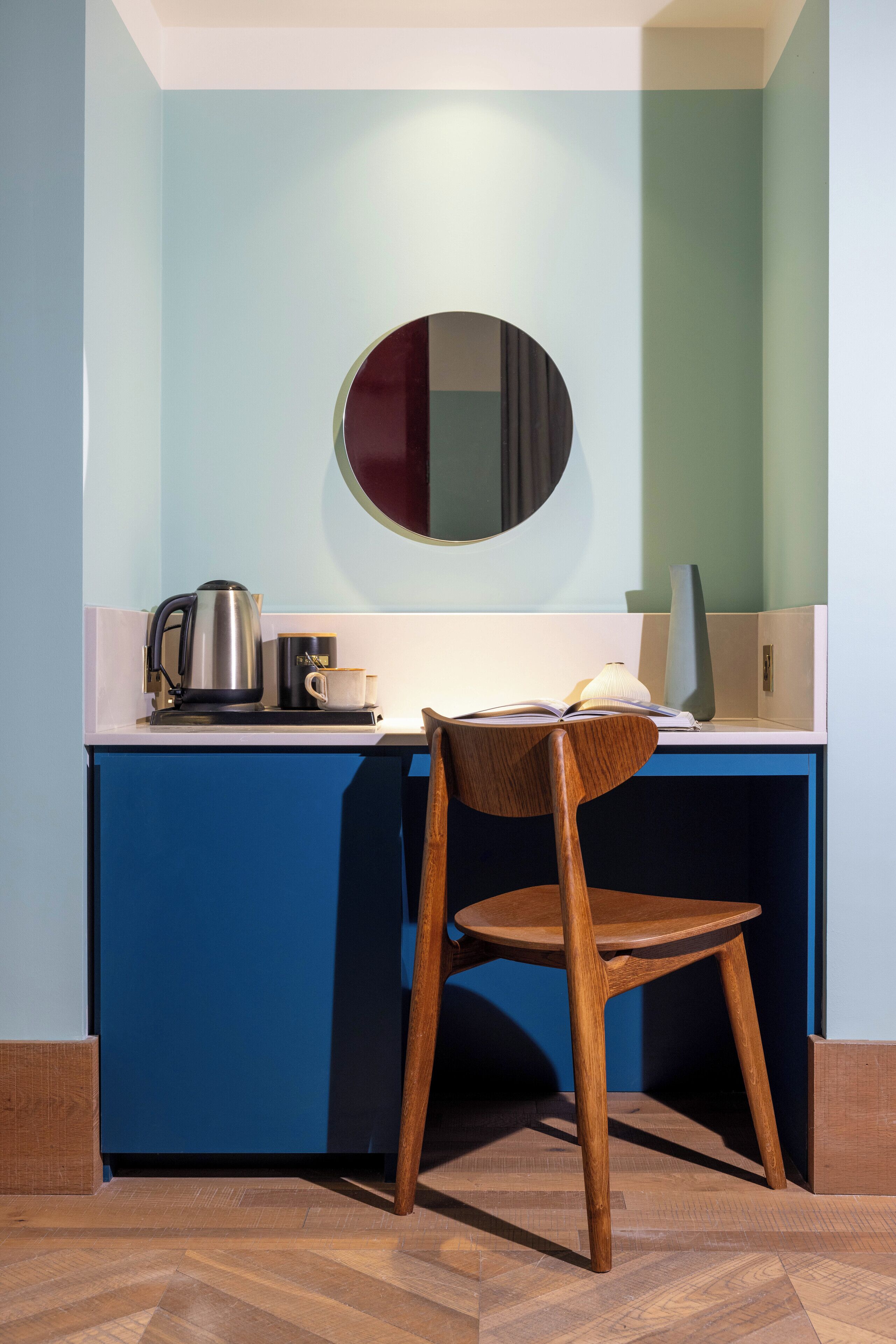 A small, modern kitchenette with a blue cabinet, a wooden chair, a kettle, a mug, and a mirror on the wall.