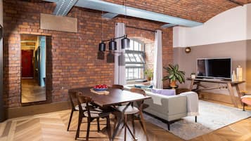 A spacious living room with a brick wall, a dining area with a wooden table and chairs, a sofa, a TV, and a potted plant.