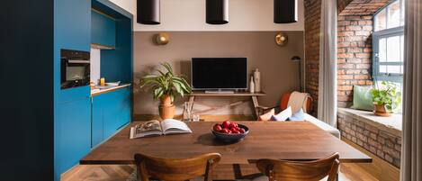 A modern kitchen with blue cabinets, a wooden dining table, and a TV in the background.
