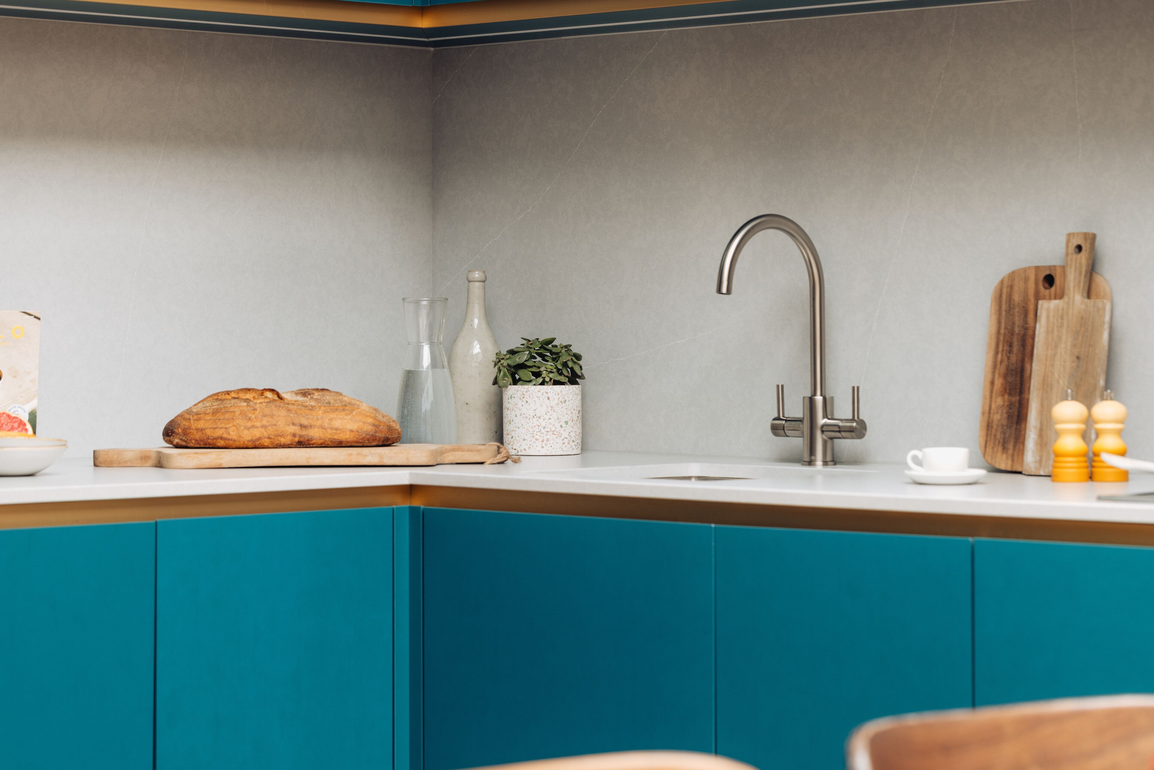Una cocina con gabinetes azul verdoso, un fregadero de acero inoxidable, una tabla de cortar de madera, una barra de pan, una planta pequeña y una tabla de cortar de madera.