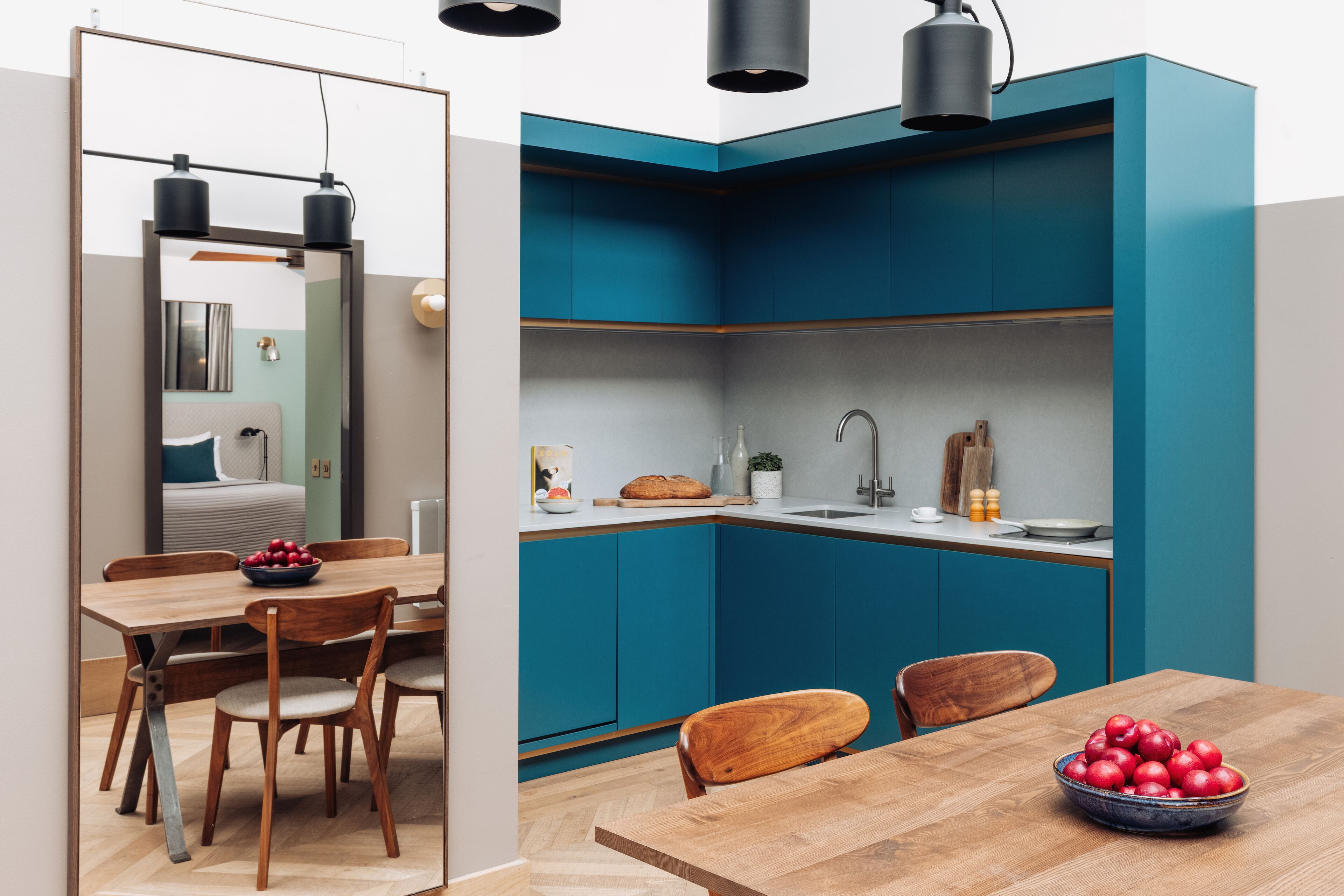A compact kitchen with teal cabinets, a wooden dining table with chairs, and a bowl of apples on the table.