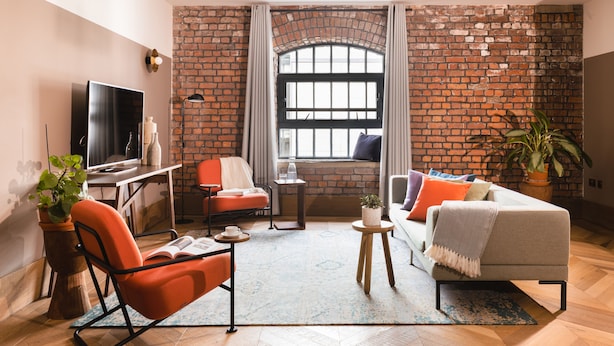 A modern living room with a brick wall, a large window, a sofa, a chair, a TV, and a small table with a plant. - Native Manchester (Manchester)