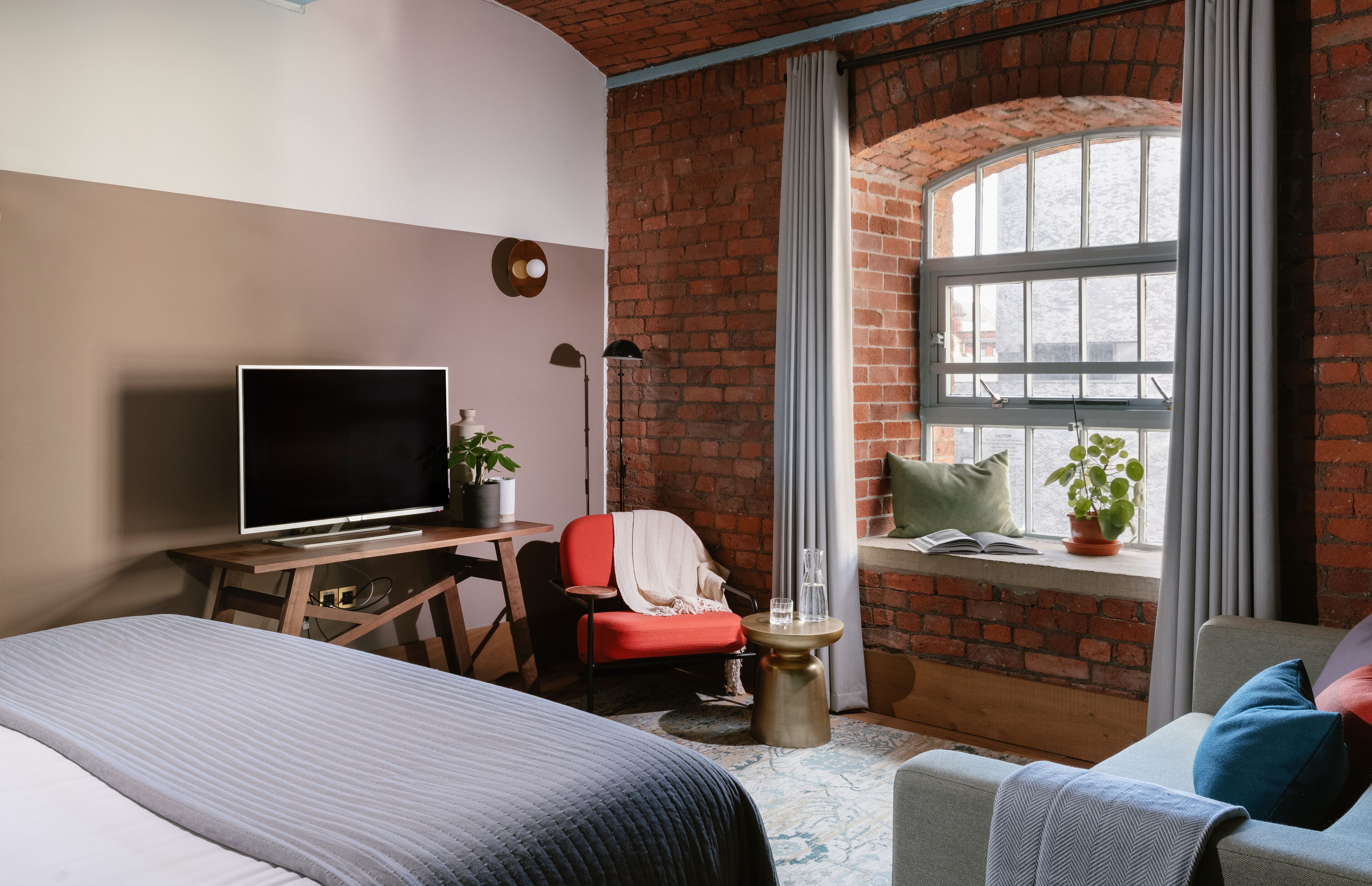 Ein Zimmer mit Backsteinwand, großem Fenster, einem Bett, einem Schreibtisch mit Fernseher und einem Sessel.