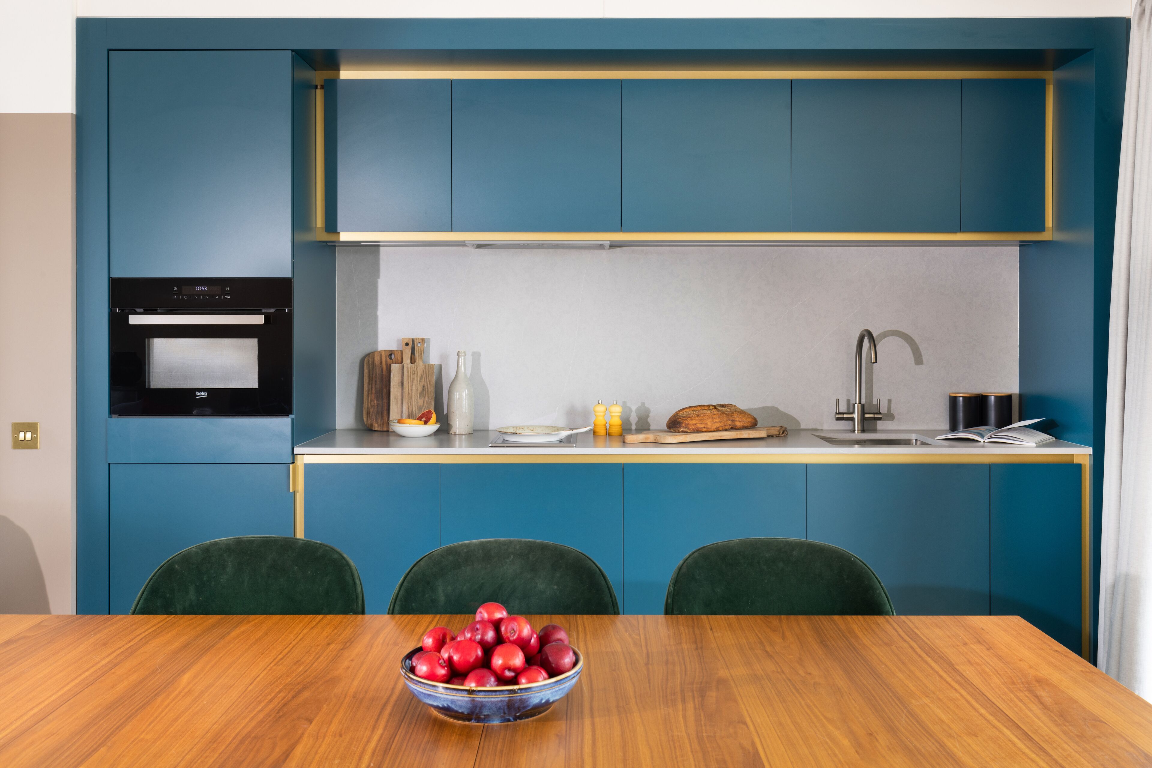 Una cocina moderna con armarios azules, horno integrado y comedor con mesa de madera y sillas verdes.