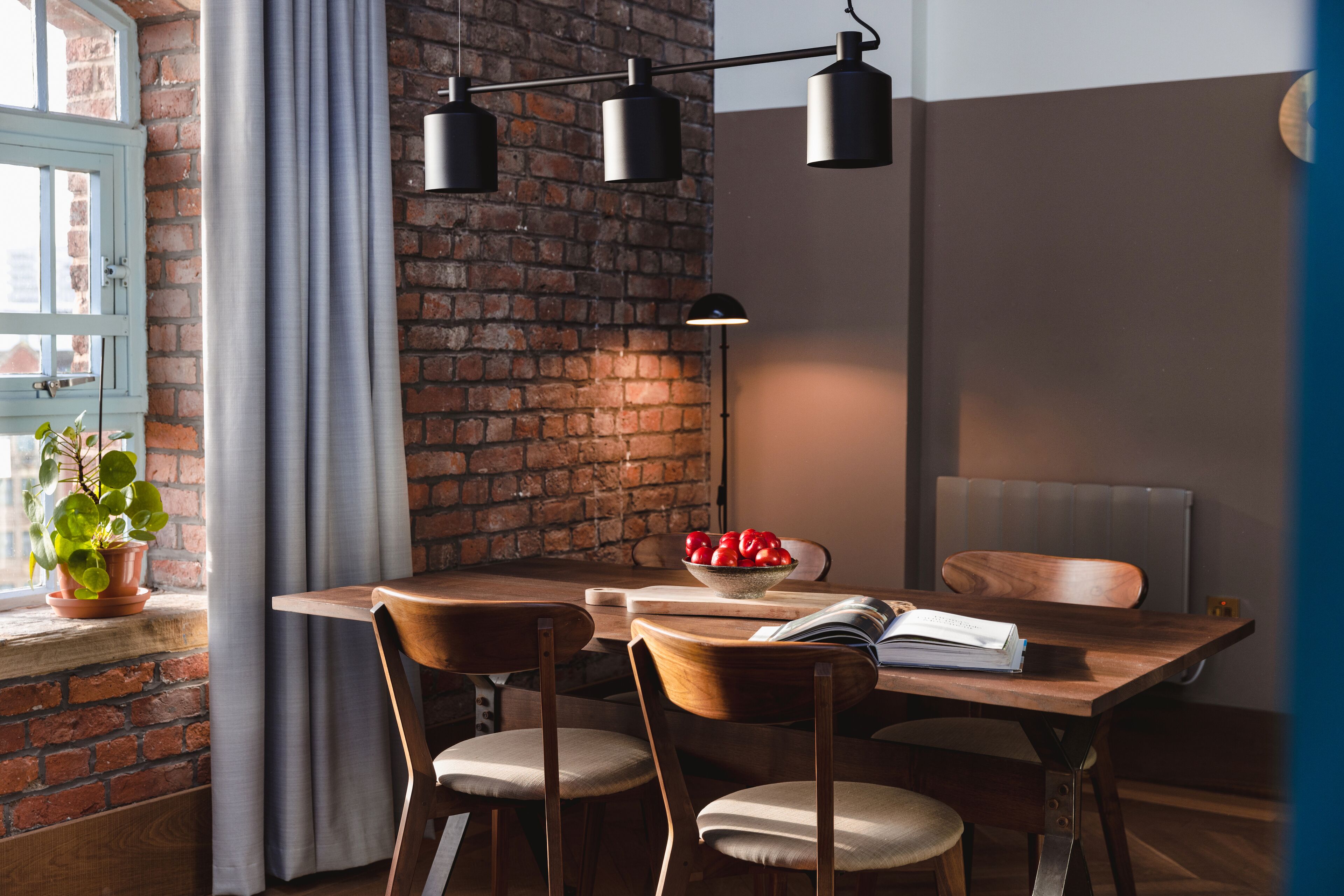 Un coin repas avec un mur en briques, une table en bois et des chaises. On peut voir un saladier de fruits sur la table et une plante sur le rebord de la fenêtre.