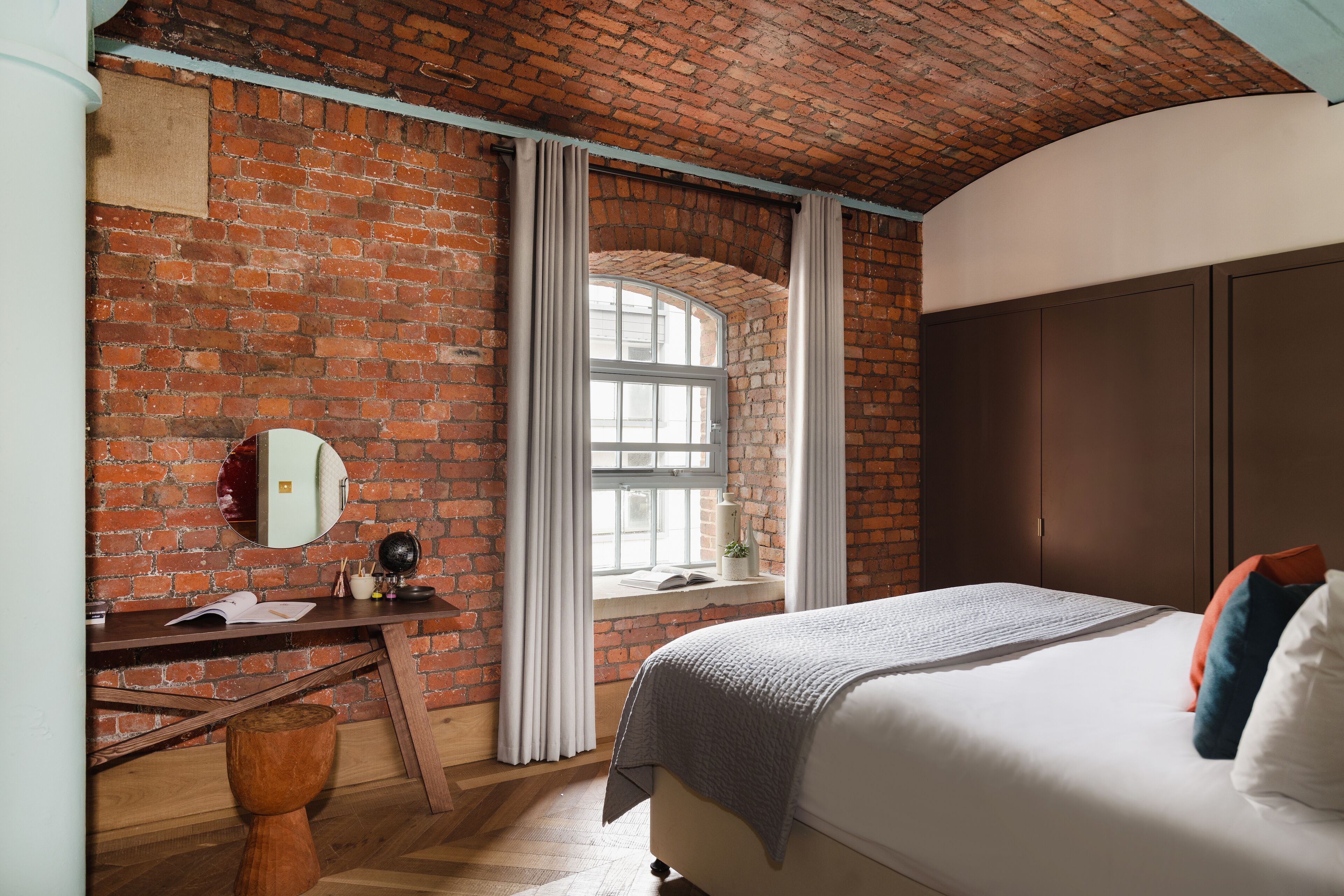 A bedroom with a brick wall, a wooden desk, a bed with pillows, and a window with curtains.