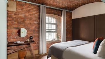 A bedroom with a brick wall, a wooden desk, a bed with pillows, and a window with curtains.