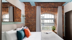 A hotel room with a brick wall, a large bed with white and teal pillows, a potted plant, and a window with curtains. - Native Manchester (Manchester)