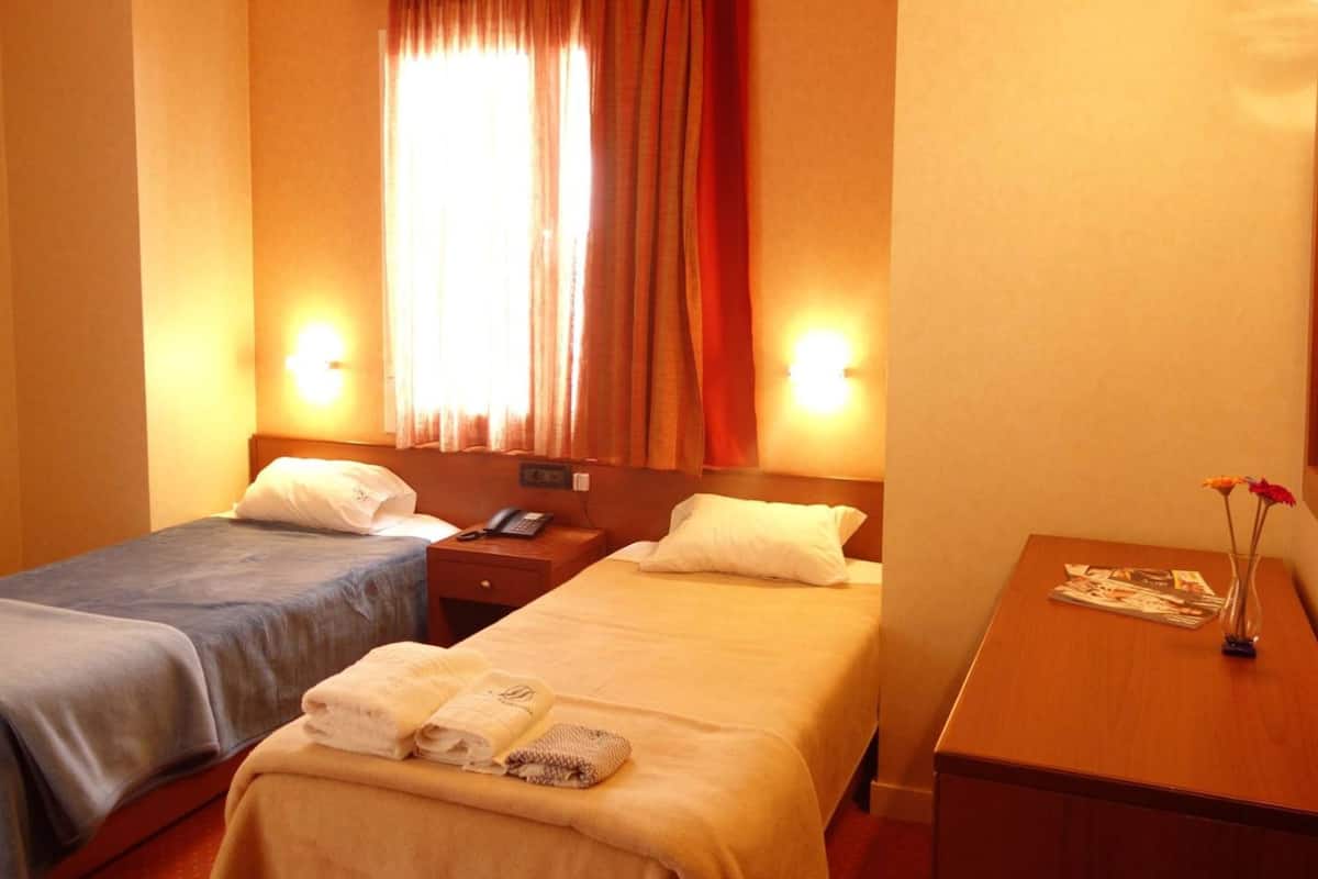 Hotellihuone, jossa on kaksi sänkyä, yöpöytä puhelimella, työpöytä lampun kanssa ja ikkuna, jossa on verhot.