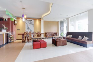 Amplia sala de estar con sofá, reposapiés, sillas de madera, una mesa de comedor y una zona de cocina con refrigerador y una planta.