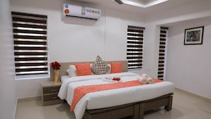 Une chambre moderne avec un grand lit, deux fenêtres équipées de stores, une table de chevet ornée d’un vase de fleurs et un appareil de climatisation.