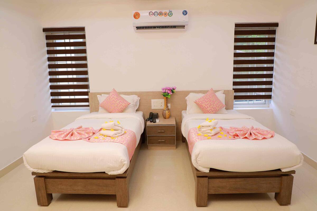 A hotel room with two beds, a bedside table with a phone and a vase of flowers, and an air conditioning unit above the beds.