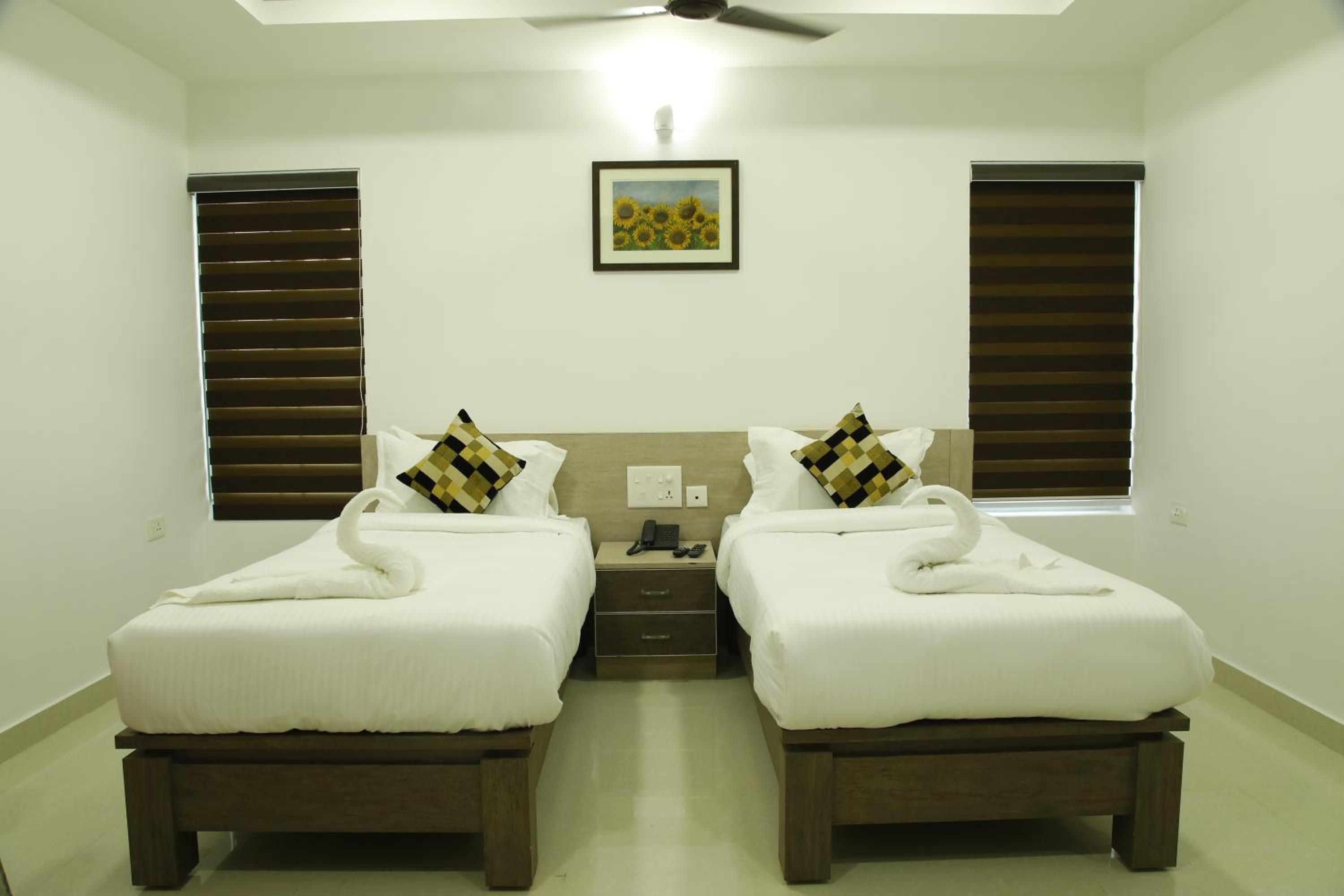 A hotel room with two beds, each with a towel and pillow, a bedside table, a ceiling fan, and a framed picture on the wall.