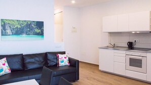 Ein modernes Wohnzimmer mit einer schwarzen Ledersofa, einem kleinen Tisch und einem farbenfrohen Kissen. Angrenzend an das Wohnzimmer befindet sich eine Küchenzeile mit weißen Schränken und einem eingebauten Backofen.