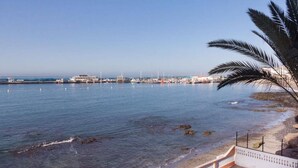 Blick auf die Küste mit blauem Himmel, Sandstrand und einem Hafen im Hintergrund mit Booten und Gebäuden.