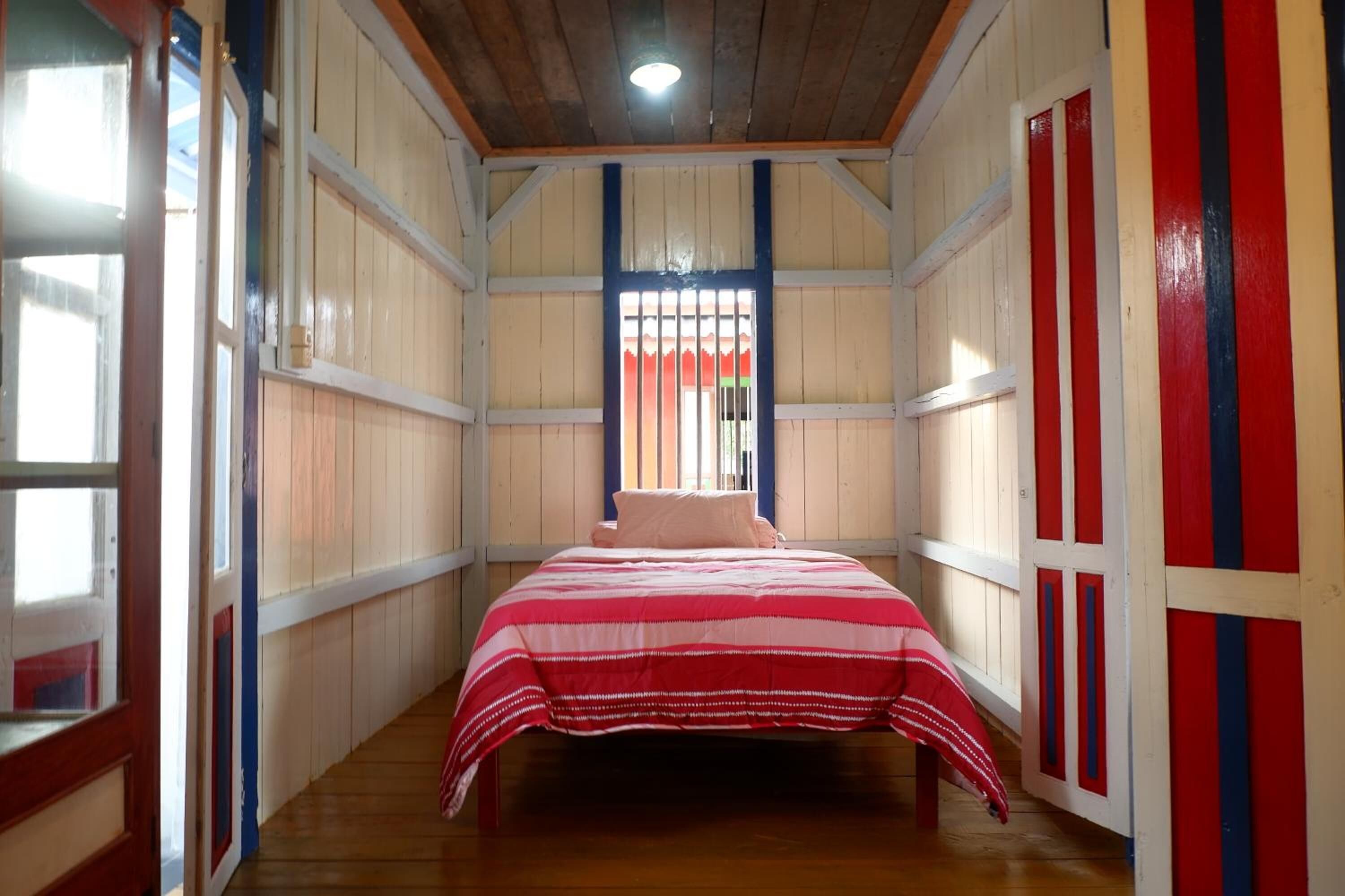 A narrow room with a bed, wooden walls, a window with bars, and a door with red and blue stripes.