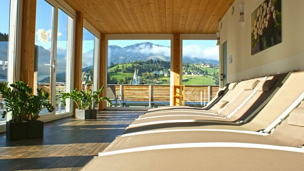 Una habitación iluminada por el sol con grandes ventanales, vigas de madera y sillones orientados hacia una vista panorámica de la montaña.