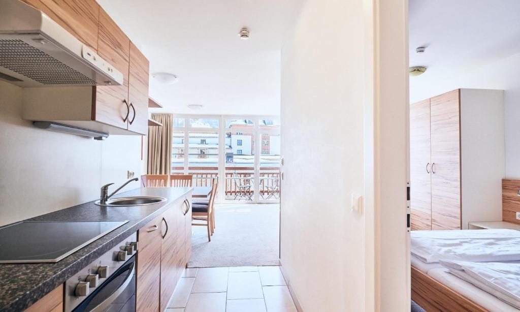 Een kleine keuken met een fornuis, wastafel en kastjes, die uitkomt op een balkon met buitenmeubels.