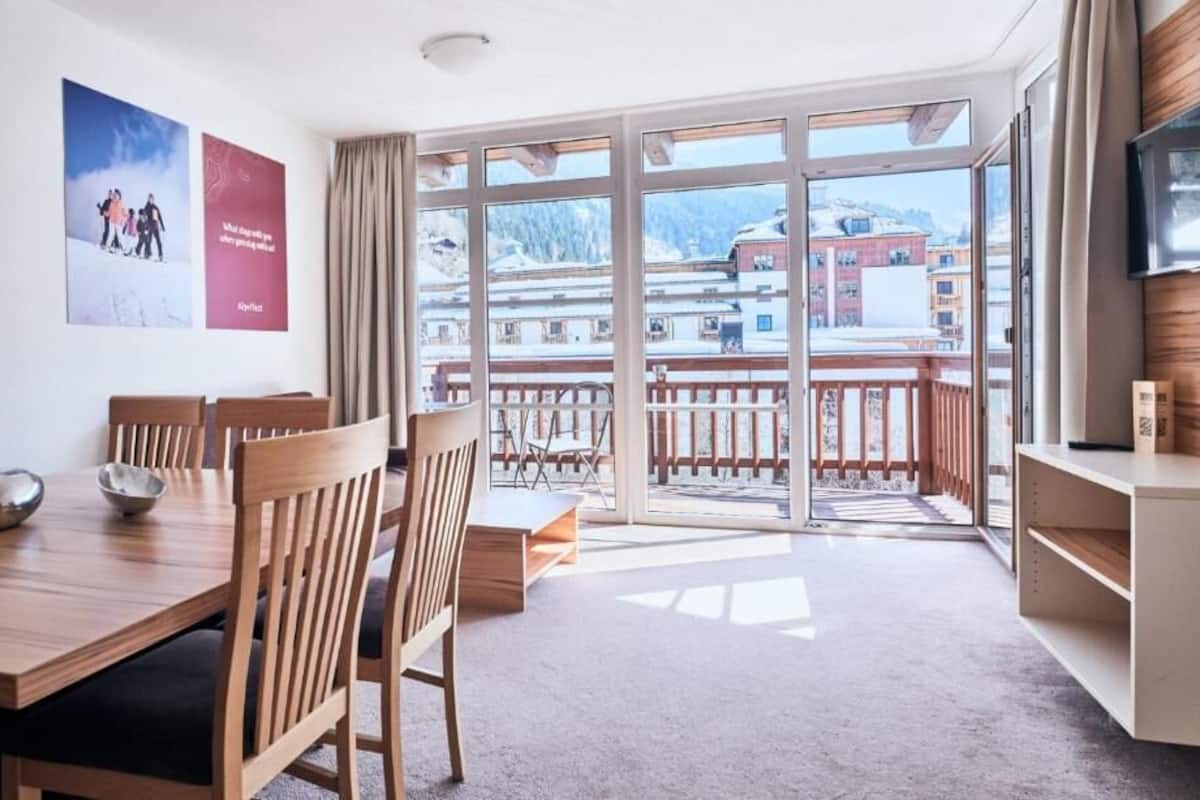 Quarto de hotel com mesa de jantar de madeira, cadeiras, televisão e varanda com vista para edifícios e montanhas.