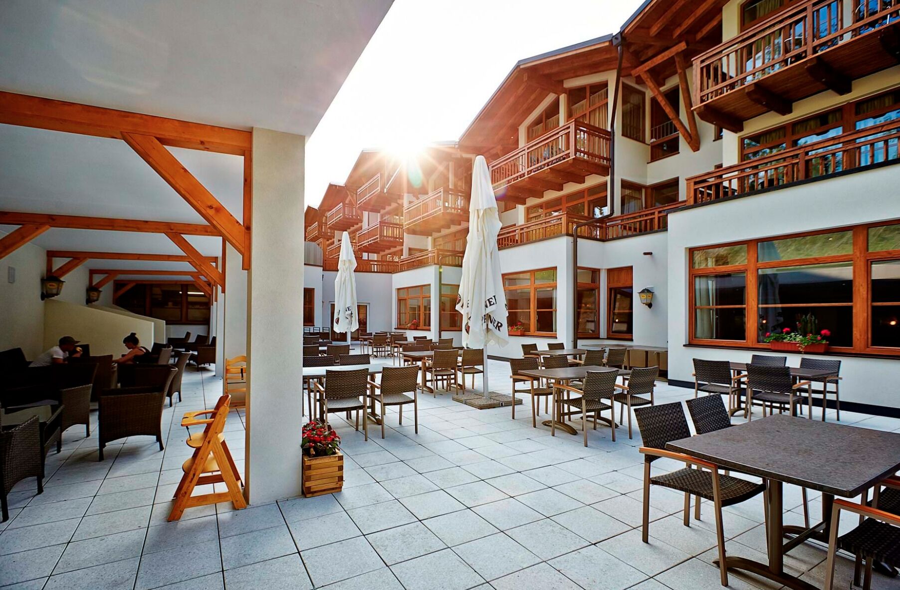 A spacious outdoor patio with multiple tables and chairs, wooden beams, and a view of a multi-story building with balconies.