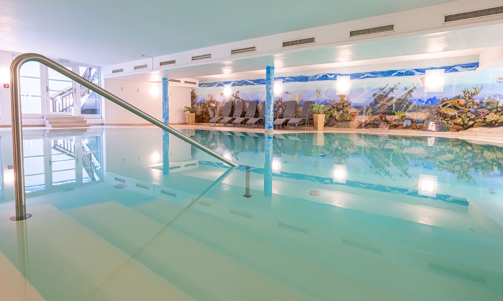 An indoor swimming pool with a clear glass floor, a metal handrail, and a mural depicting a marine scene on the wall.