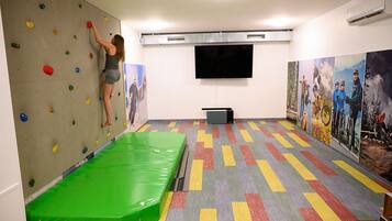 Eine Kletterwand mit bunten Griffen, einer grünen Stoßmatte und einem Flachbildfernseher an der Wand.