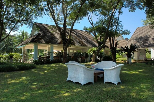 A covered outdoor seating area with wicker furniture, surrounded by lush greenery and palm trees.