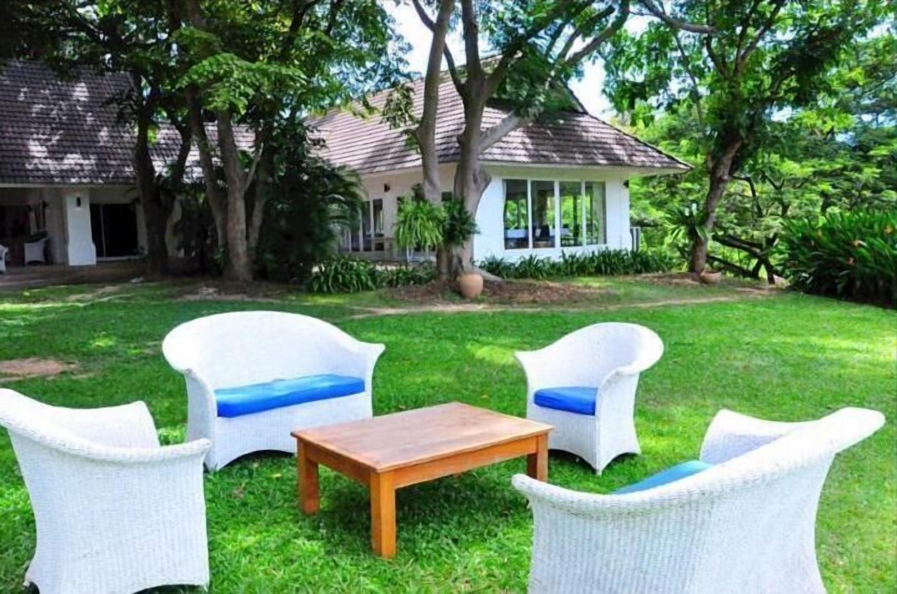 A house with a tiled roof, surrounded by lush greenery and a well-maintained lawn. There are two white wicker chairs with blue cushions and a wooden table in the foreground.
