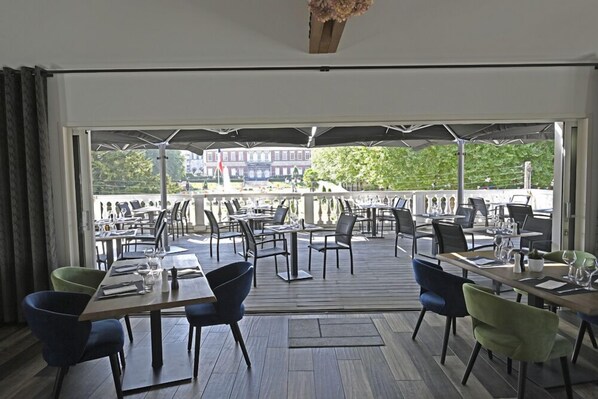 An indoor dining area with tables and chairs, large windows, and a view of a building and greenery outside. - Manoir de l'évêché et Spa (Lisieux)