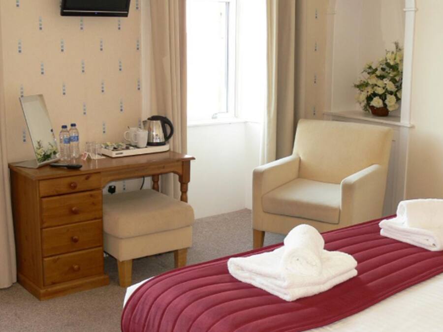 Habitación de hotel con tocador de madera, una silla, cama con colcha roja y ventana con cortinas.