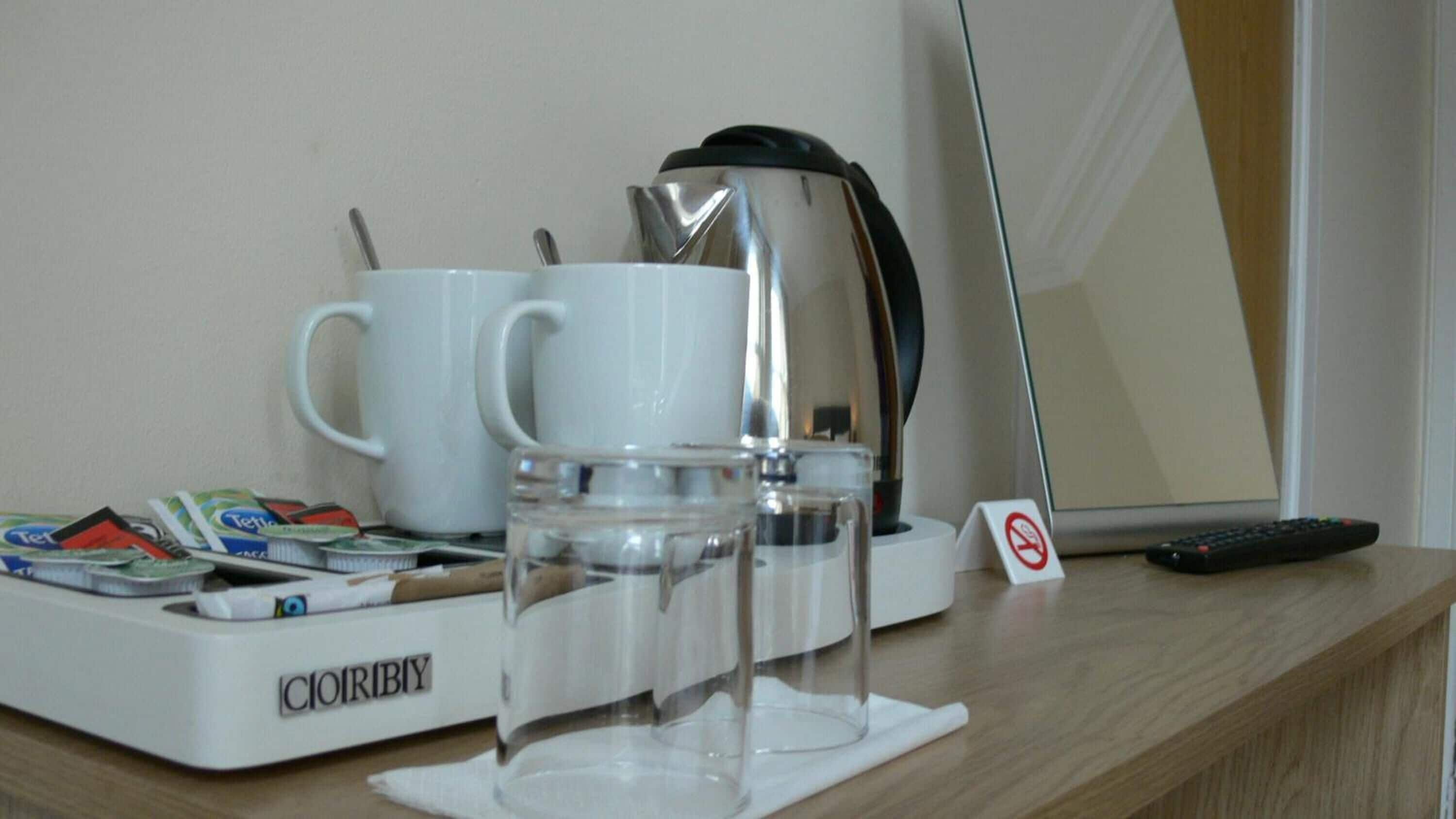 A hotel room with a minibar, two white mugs, a coffee maker, a glass jar, a remote control, and a no-smoking sign.