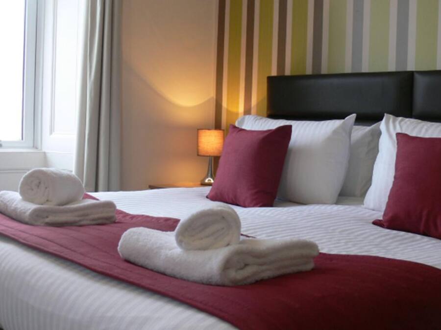 Un lit bien fait, avec des draps blancs, un plaid rouge et deux serviettes enroulées.
