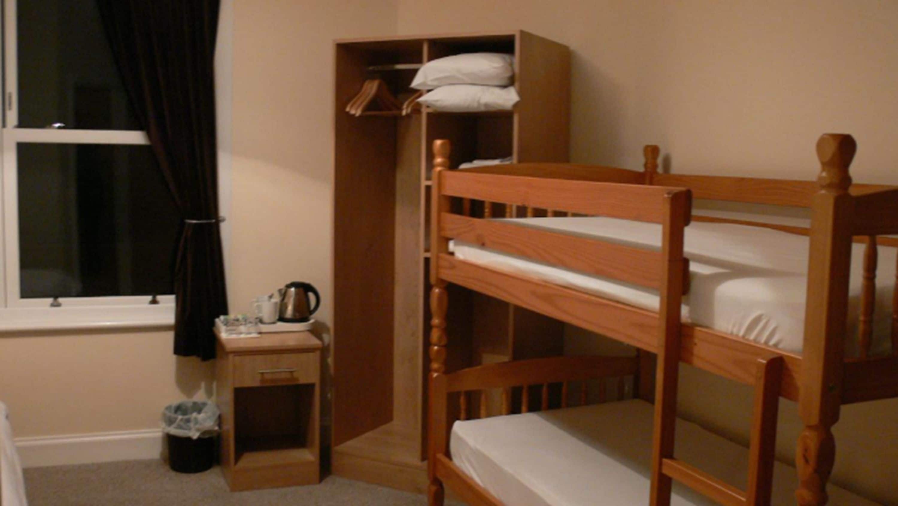 A bunk bed room with a small bedside table, a shelf with a kettle, and a window with curtains.
