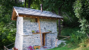 Una cabaña de piedra con techo inclinado, vigas de madera y un pequeño porche con macetas y flores.