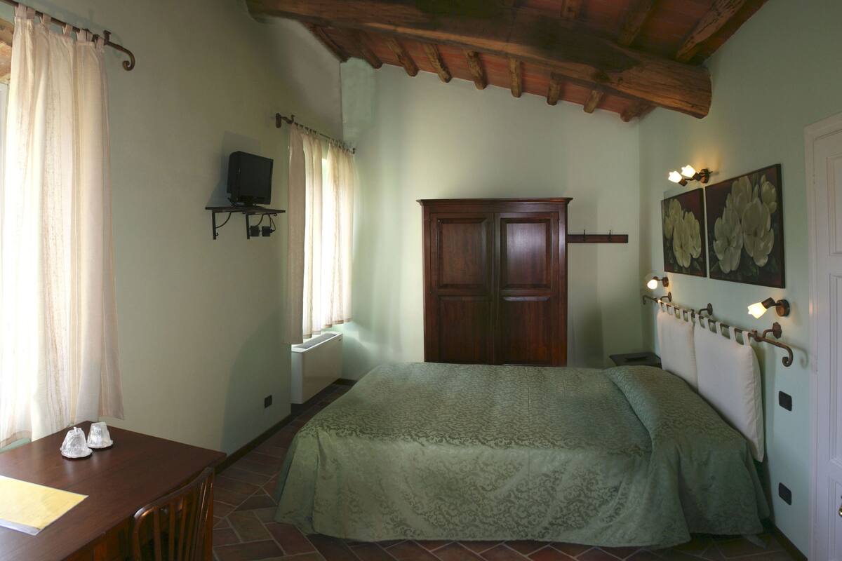 Un dormitorio con cama, un armario de madera, una pequeña mesa con sillas, un televisor sobre un soporte y una ventana con cortinas.