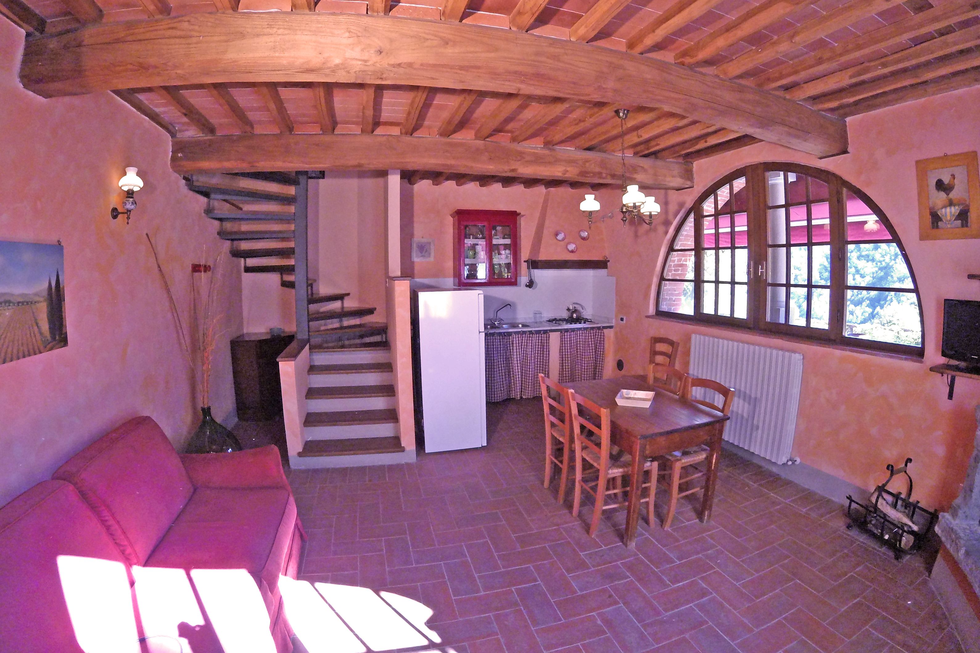 Um interior acolhedor com uma escada de madeira, uma área de refeições com mesa e cadeiras, uma cozinha com frigorífico e uma grande janela em arco.