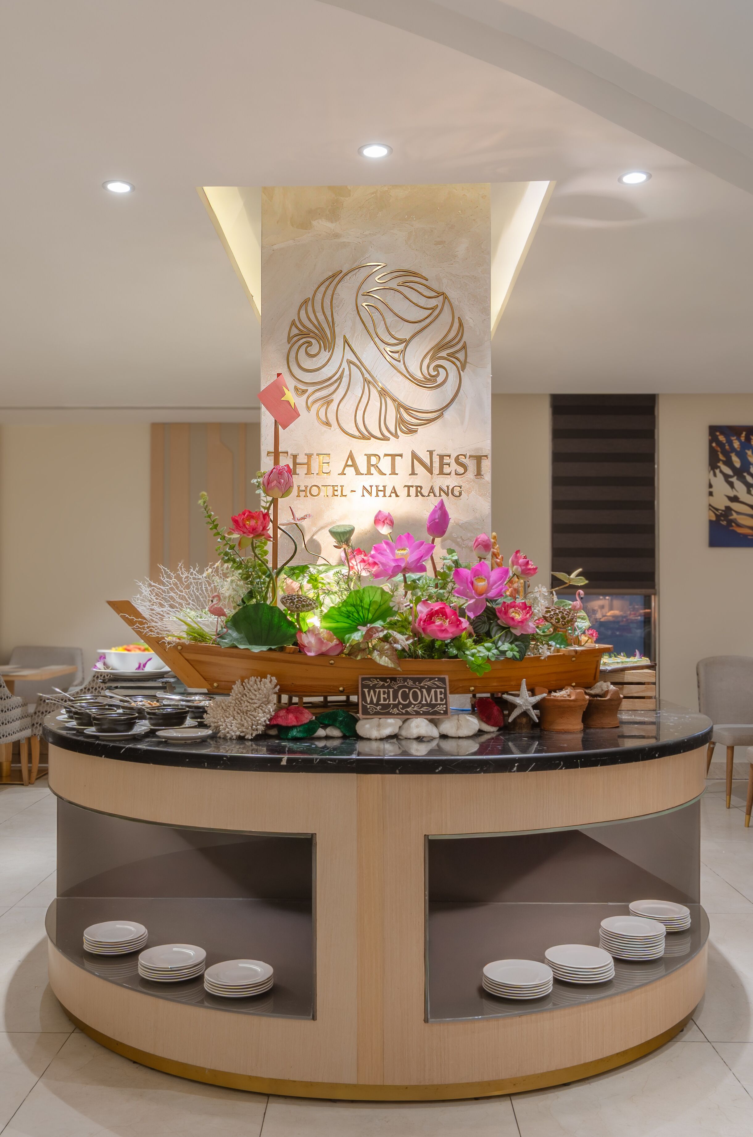 Een receptiebalie bij The Art Nest Hotel-Nha Trang, met een bloemstuk, een welkomstbord en netjes gestapelde borden.