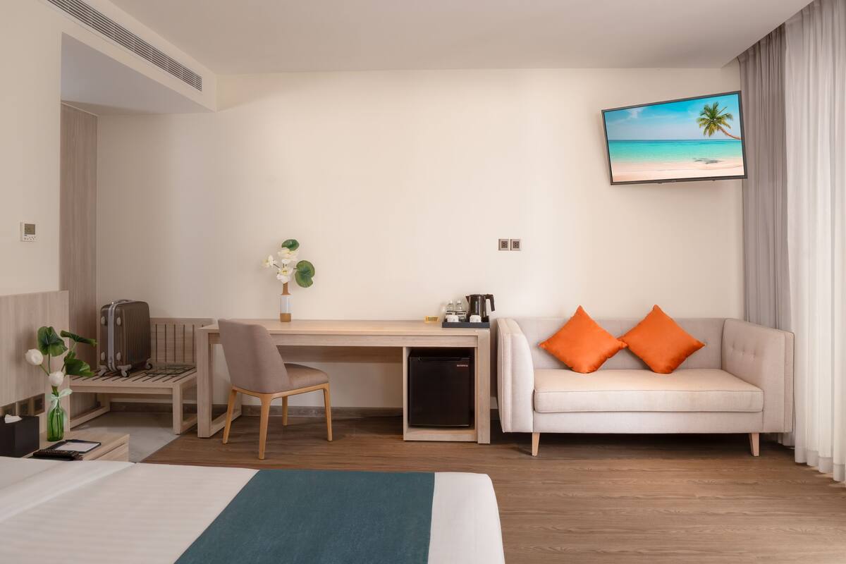 A hotel room with a bed, a desk, a chair, a sofa, and a TV displaying a beach scene.