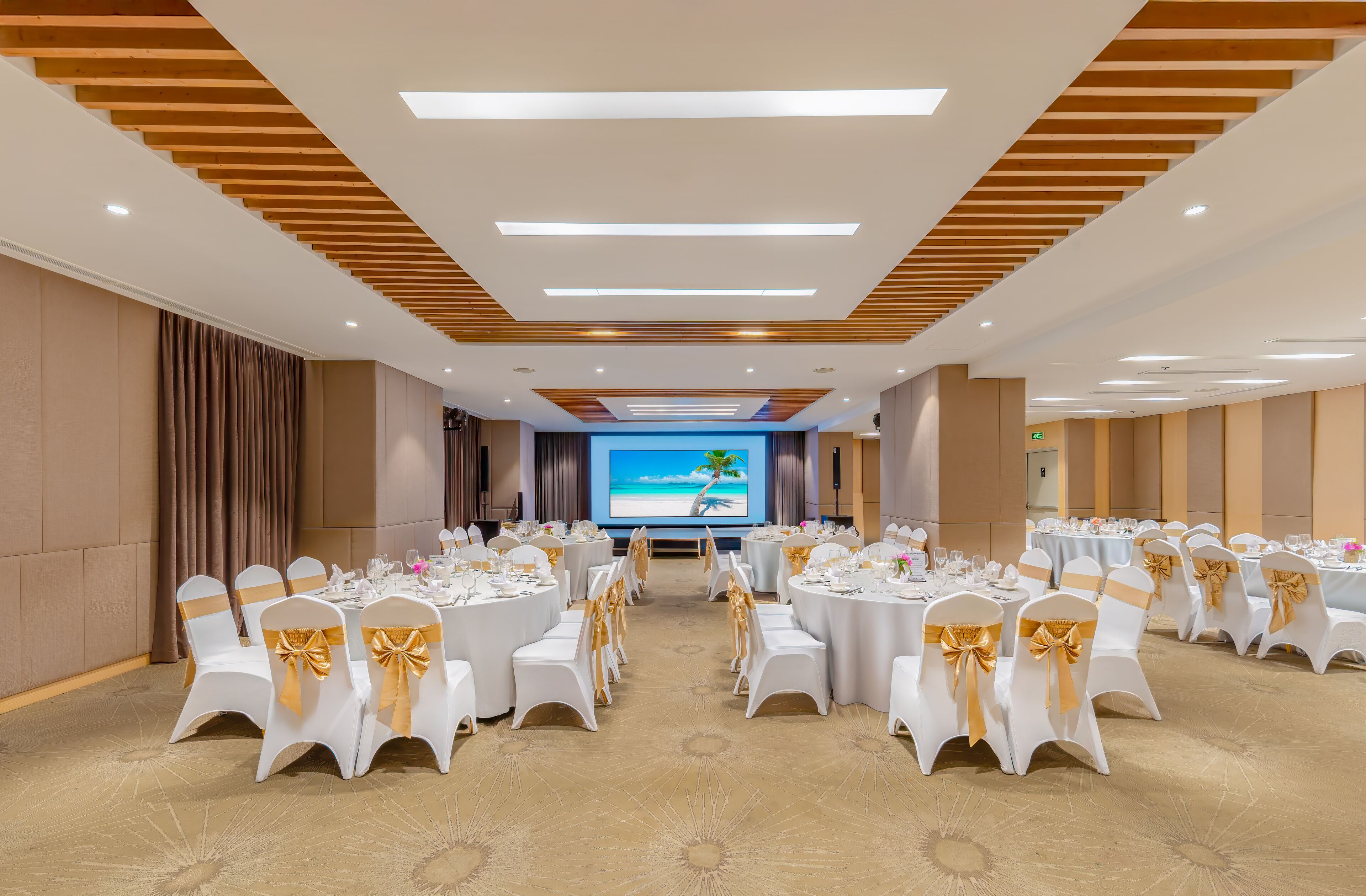 A conference room with white tables and chairs, a large screen displaying a beach scene, and a wooden ceiling design.