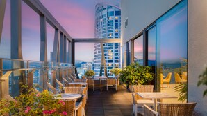 A rooftop terrace with wicker furniture, potted plants, and a view of a cityscape at dusk.