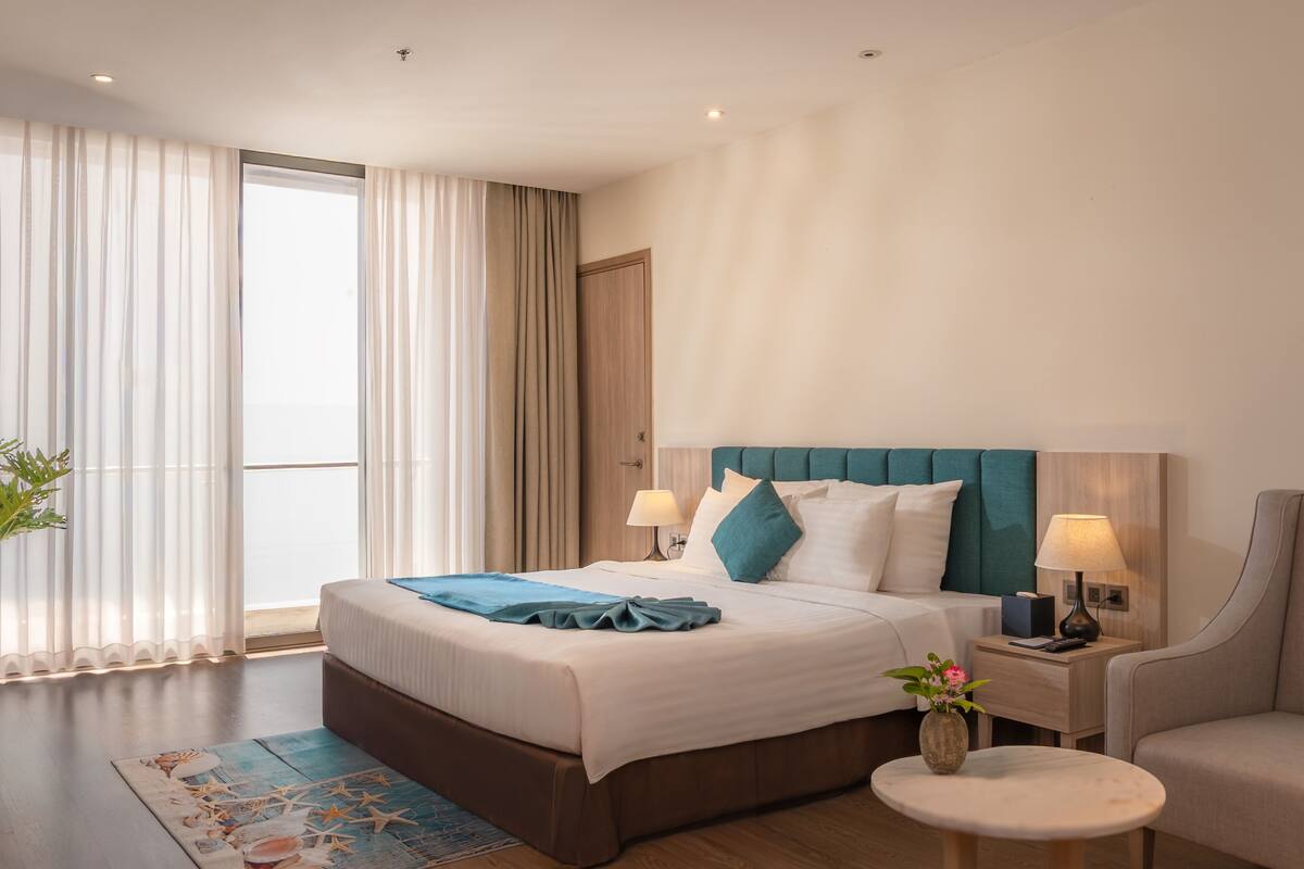 A modern hotel room with a large bed, a nightstand with a lamp, a round table, and a chair.