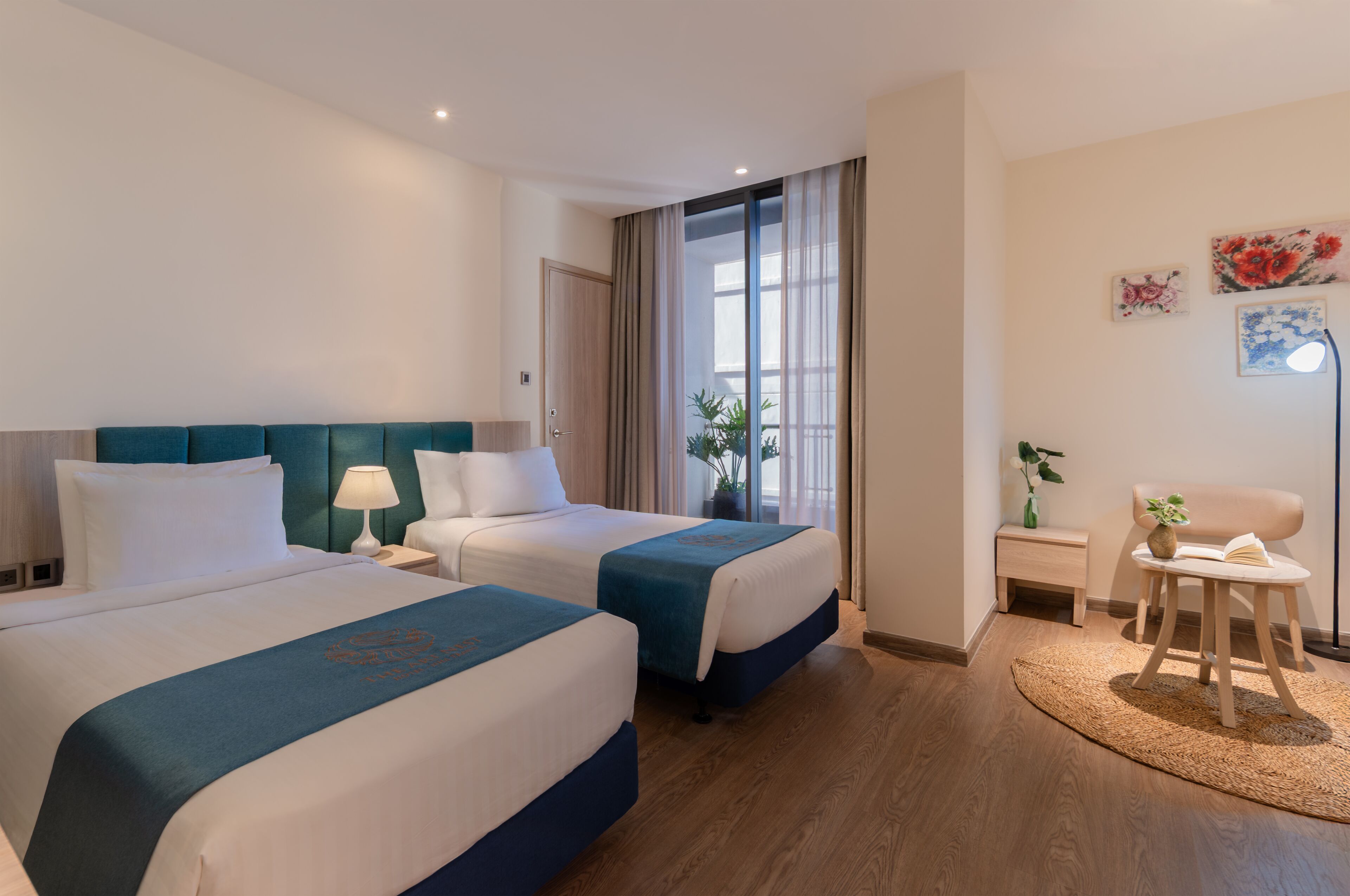 A hotel room with two beds, a small table with a lamp, a chair, and a view of the outdoors.