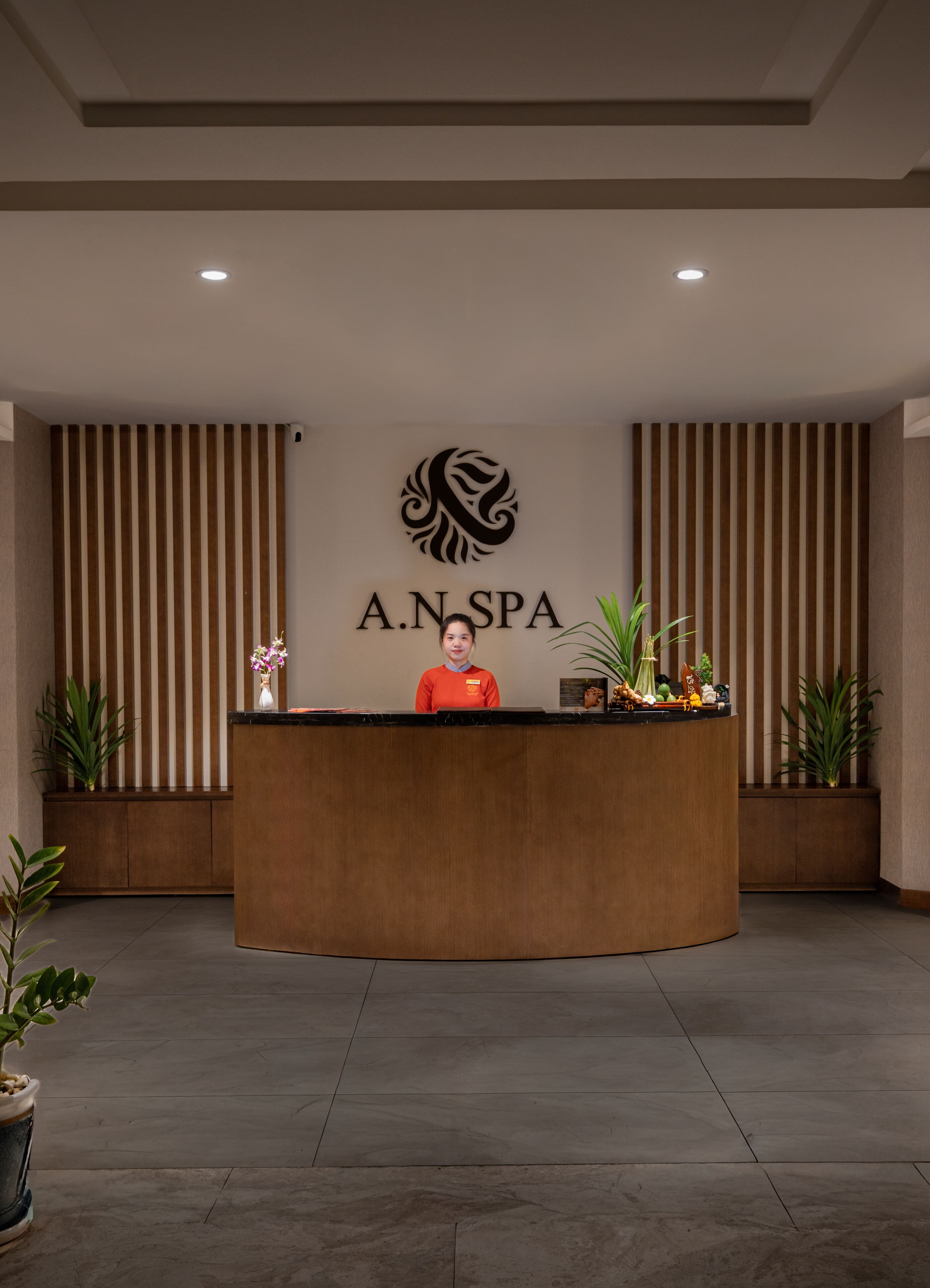 Un comptoir d’accueil orné d’un logo, une réceptionniste et des plantes en pot.