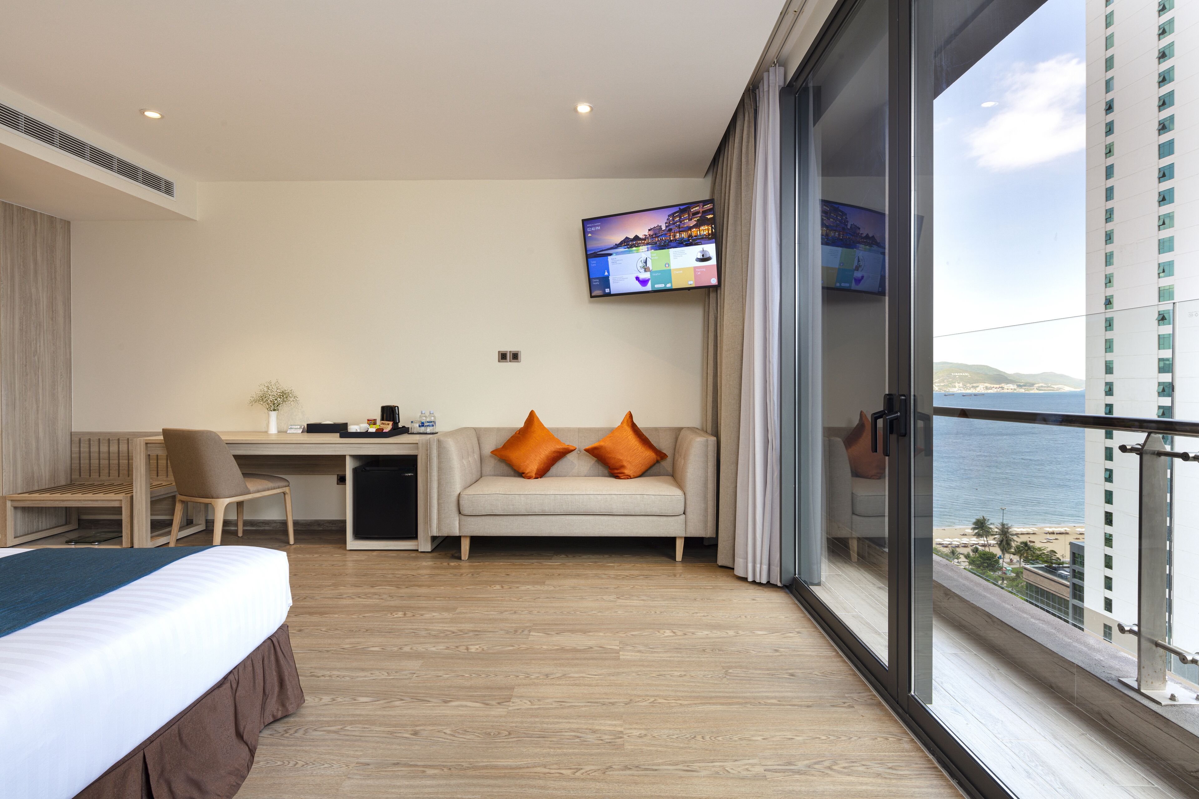 Une chambre d’hôtel comprenant un lit, un bureau, un canapé avec des coussins orange, un téléviseur fixé au mur et un balcon offrant une vue sur la mer et les bâtiments.