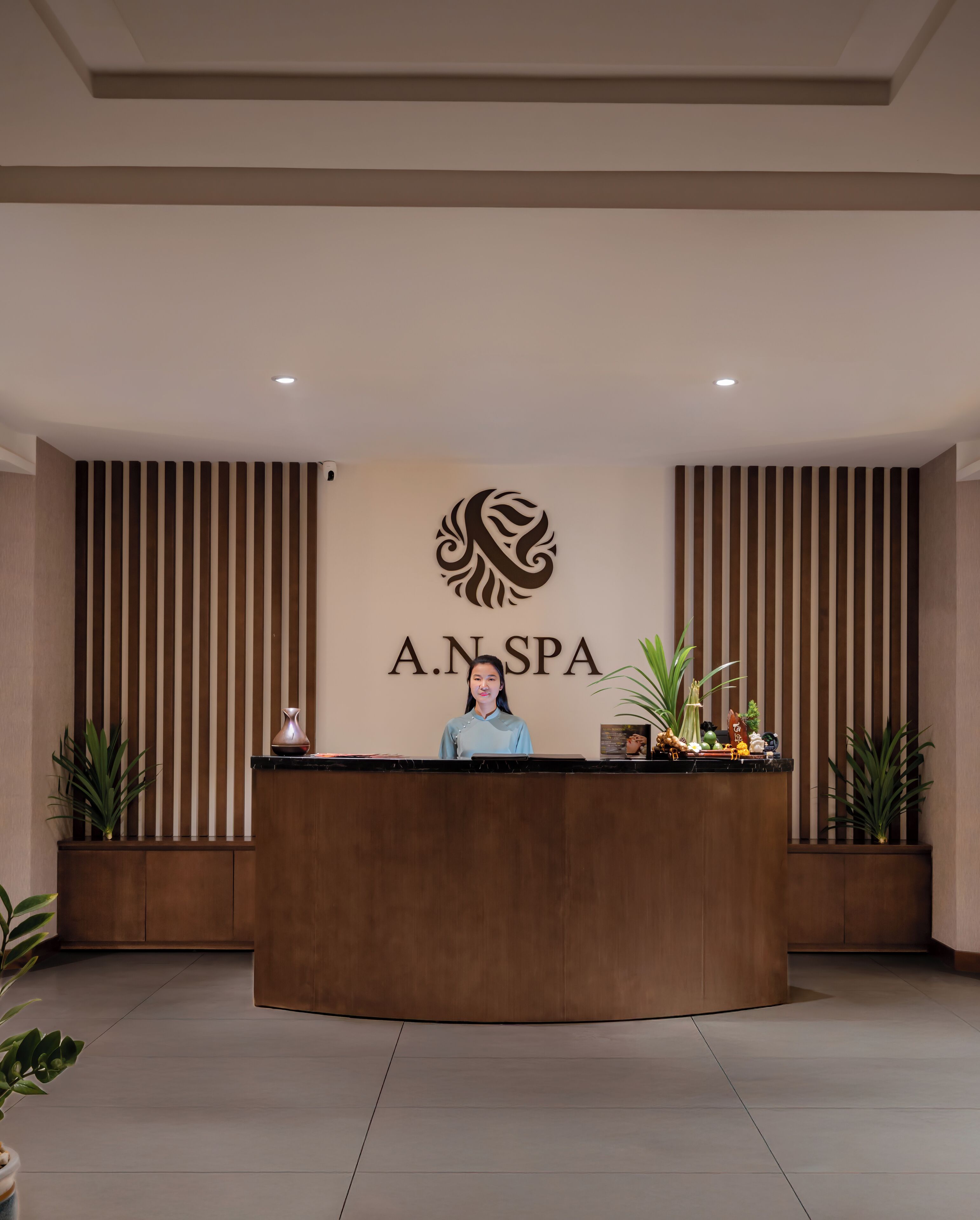 A reception desk with a logo and name "A.N.SPA" in the center, flanked by vertical wooden slats, and potted plants on either side.