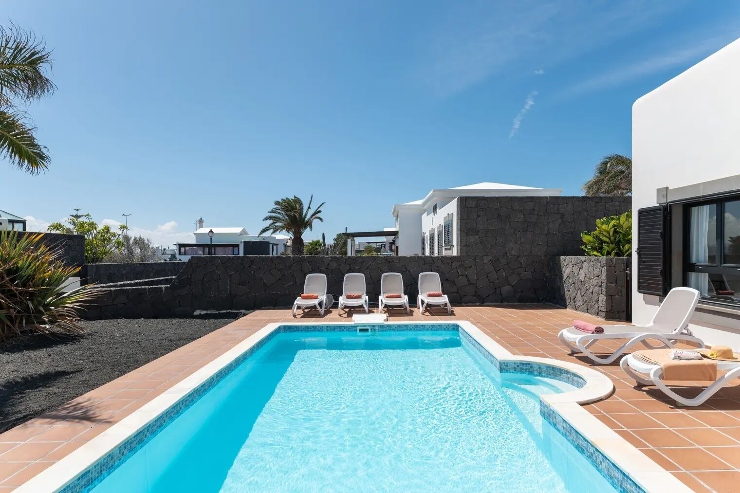 A rectangular swimming pool with clear blue water, surrounded by a tiled deck. There are four white lounge chairs with red cushions aligned along the pool's edge. The pool is adjacent to a modern building with a stone wall and a large window.