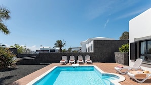 A rectangular swimming pool with clear blue water, surrounded by a tiled deck. There are four white lounge chairs with red cushions aligned along the pool's edge. The pool is adjacent to a modern building with a stone wall and a large window.