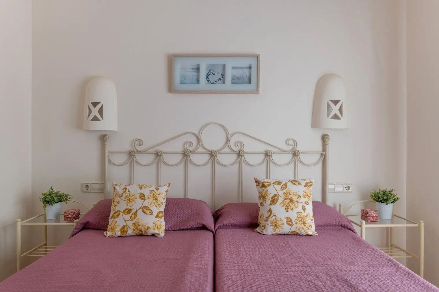 Un lit double avec une tête de lit en métal blanche, deux oreillers assortis et une couvre-lit rose. Il y a deux tables de chevet avec des lampes et de petites plantes en pot.